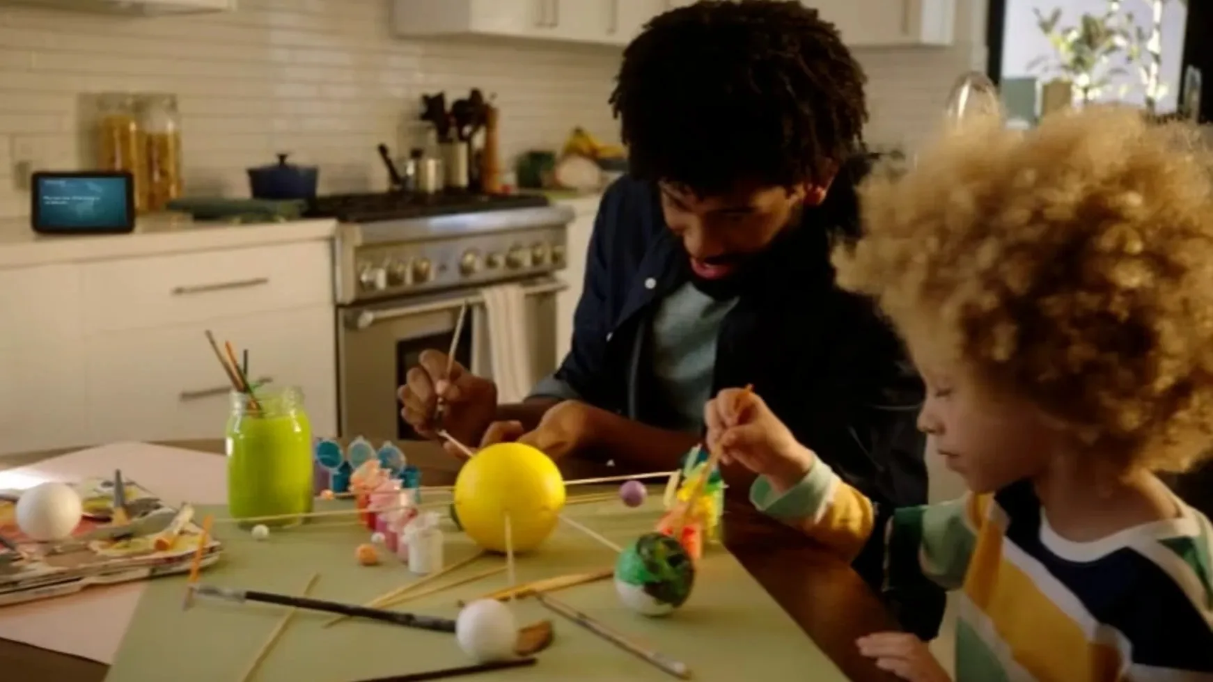 A father and his young son paint a model solar system at the kitchen table, an Echo Show on the counter behind them.
