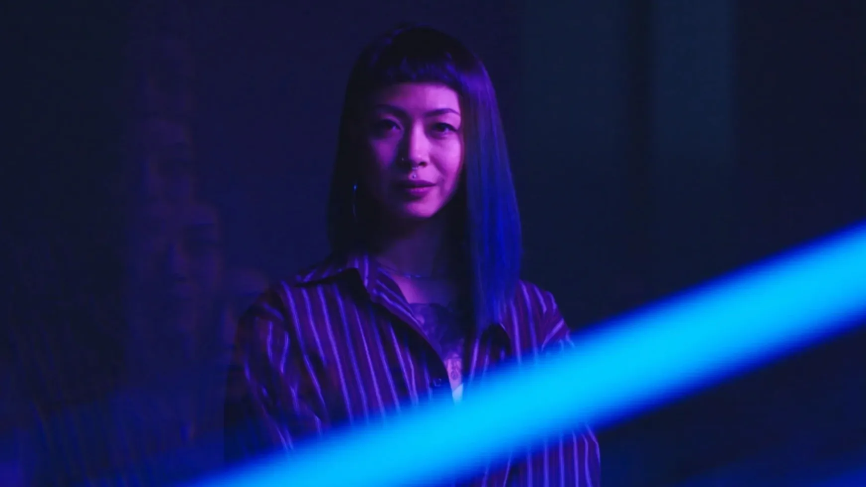 A woman with a sharp dark fringe in a striped shirt, lit violet, a beam of blue light cutting across the frame.