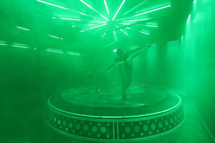 A dancer spins on a round platform in green haze under a starburst of light tubes.