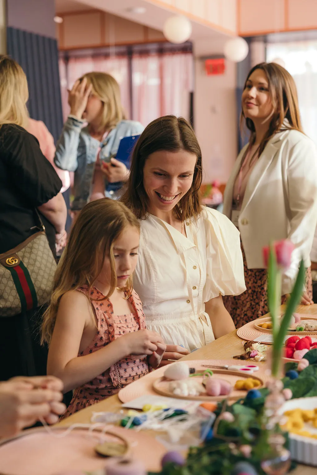 A mother smiles as her daughter threads felt shapes at a workshop table set with pink placemats.