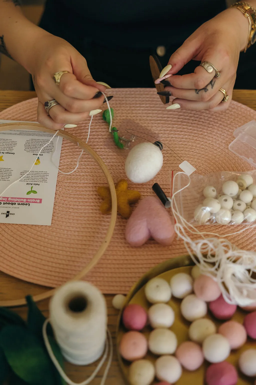 Hands thread a felt egg, star and heart onto string beside a wooden hoop and a One A Day workshop card.