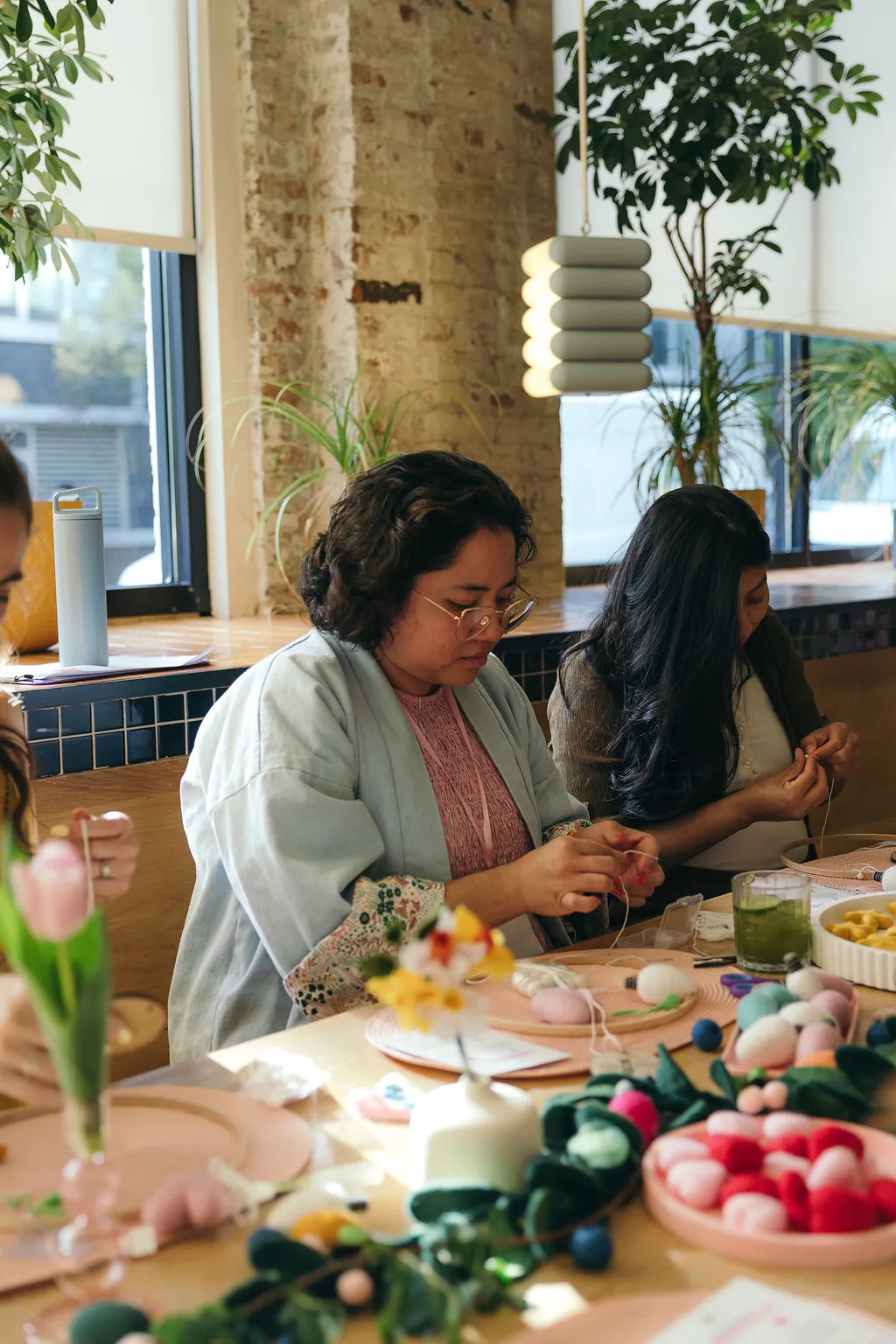 Two guests string felt mobiles at a long table in a bright room with brick and plants.