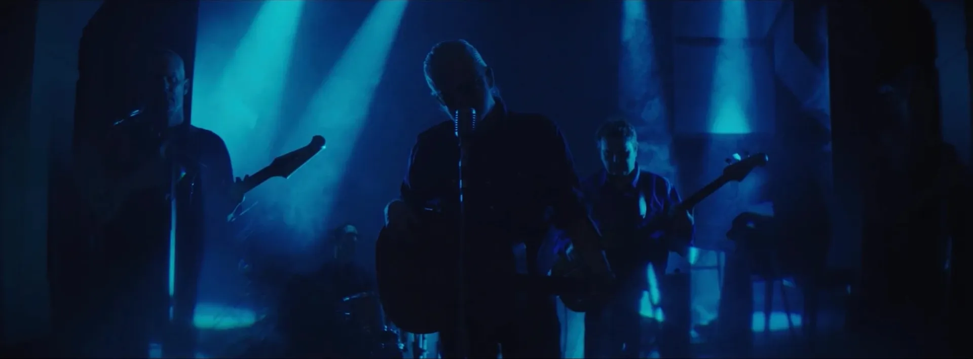 Blue Rodeo in silhouette on a smoky stage, the singer at a vintage microphone between two guitarists, under shafts of blue light.