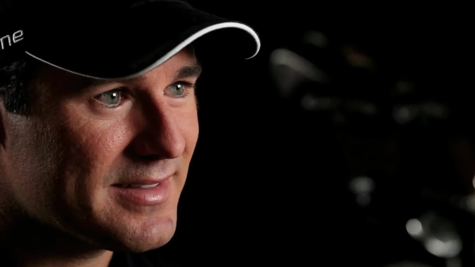 Close-up of a smiling man in a black cap, lit low against a dark room of blurred golf clubs.