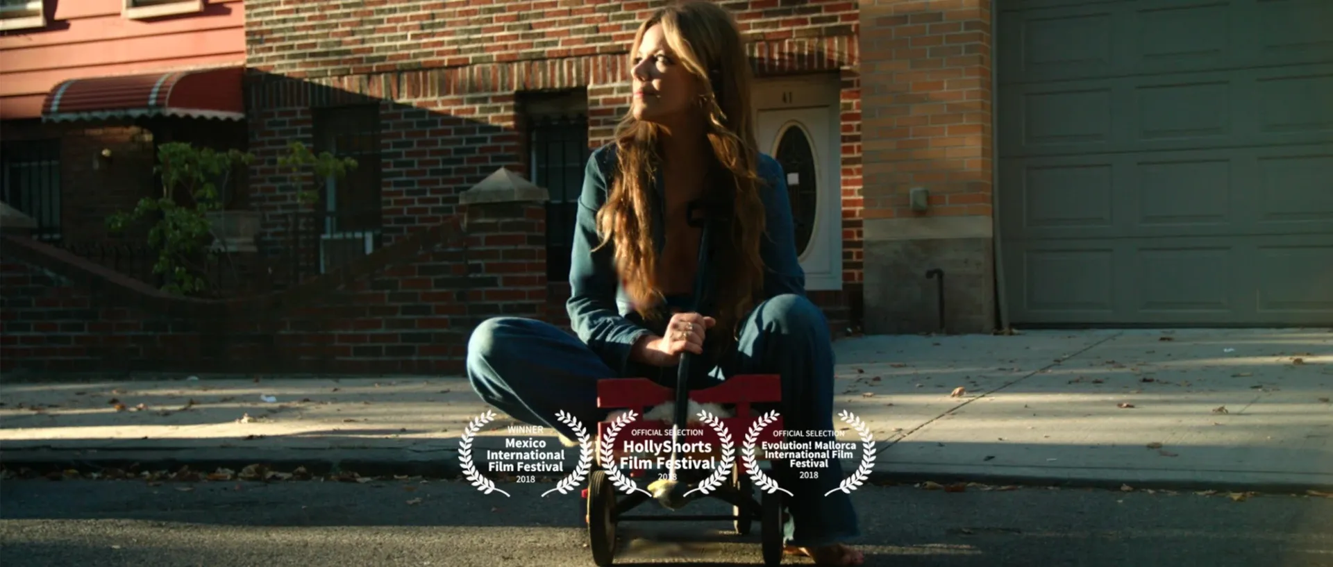 A woman in a denim jumpsuit sits cross-legged on a red wagon in a sunlit Brooklyn street, with laurels for Mexico International Film Festival (Winner, 2018), HollyShorts Film Festival and Evolution! Mallorca International Film Festival (Official Selection, 2018).