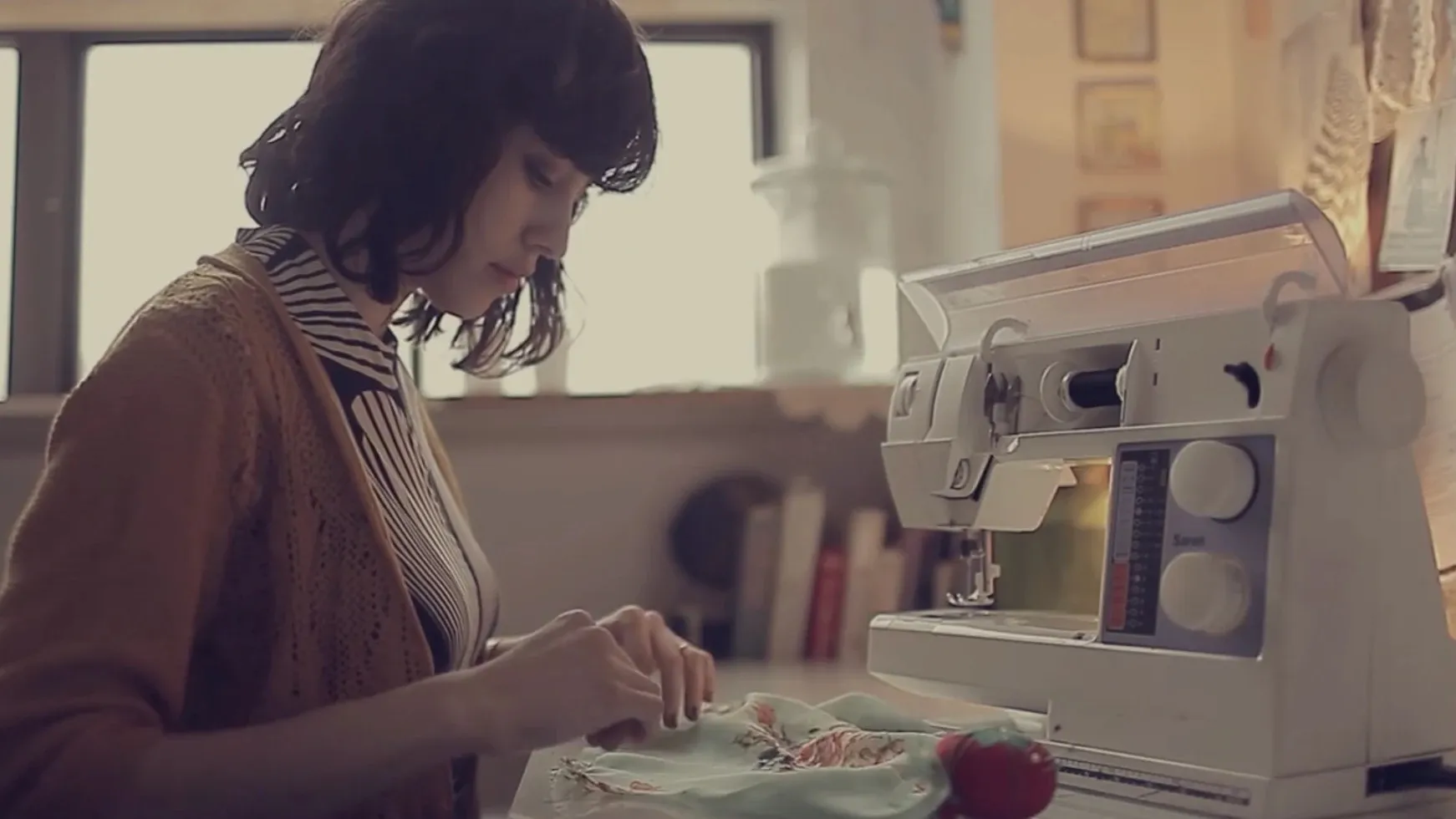 A woman in a mustard cardigan hand-stitches a floral cloth beside a sewing machine in a sunlit workroom.