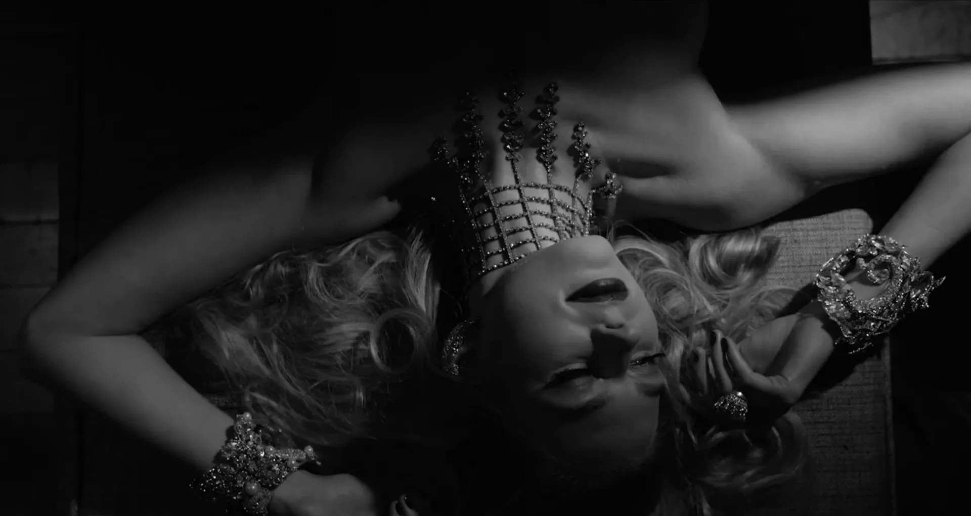 Black-and-white overhead shot of a woman lying back in a jewelled crown and crystal cuffs.