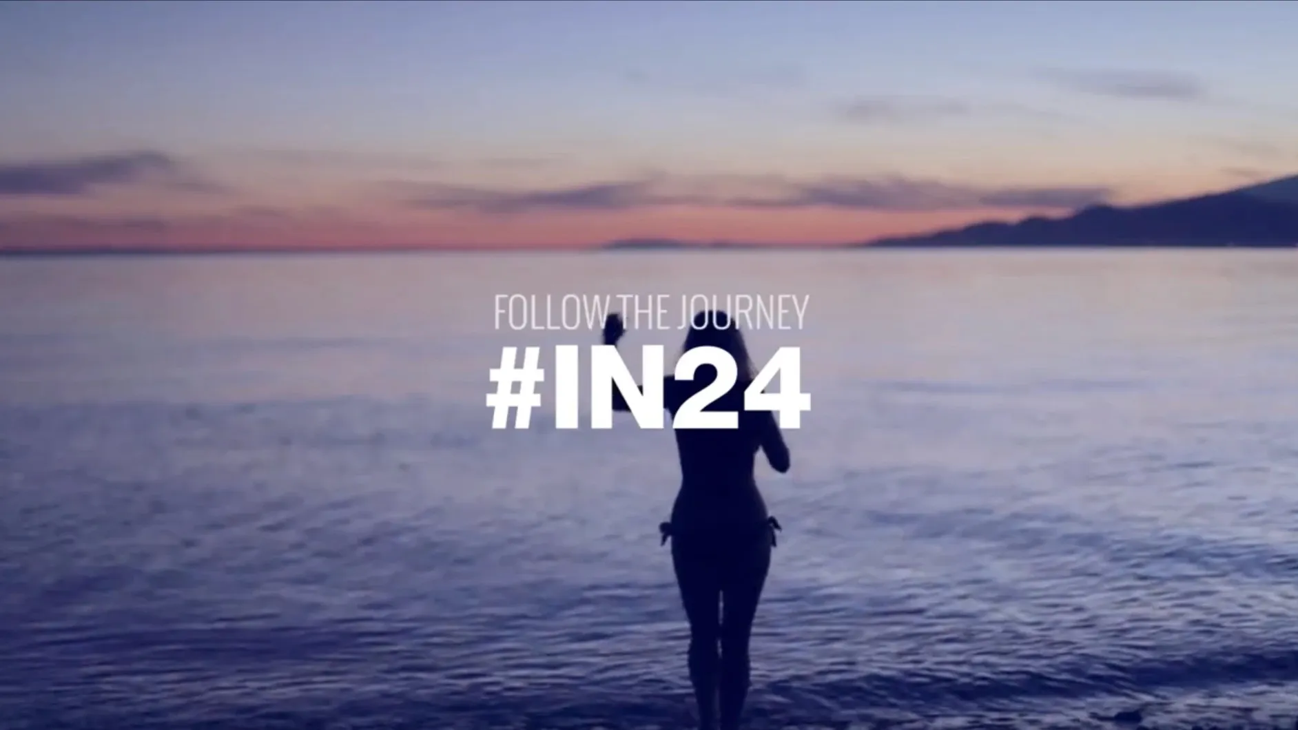 A woman in silhouette wades into the sea at dusk under a pink and violet sky, with the words Follow the journey #IN24.