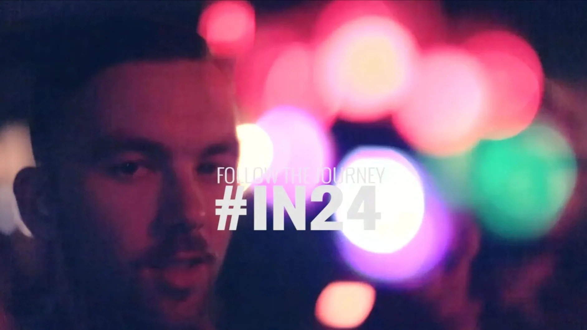 A man lit pink at night against glowing bokeh lights, with the words Follow the journey #IN24.
