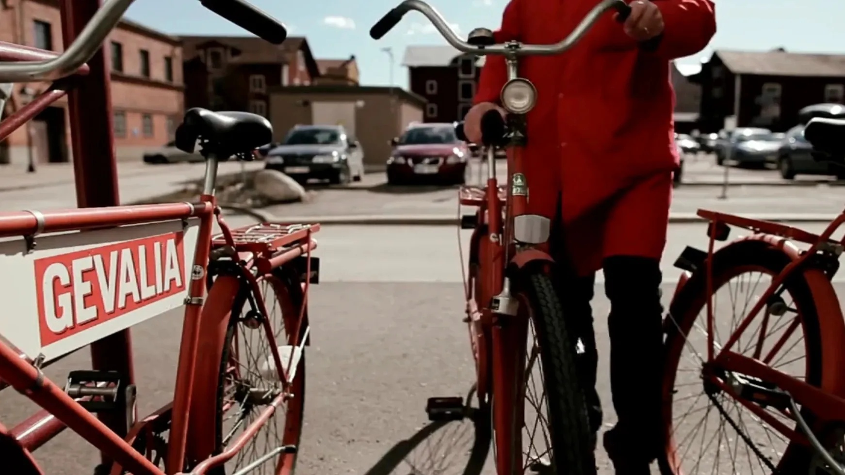 Red Gevalia delivery bikes in a town square, a man in a red Gevalia coat wheeling one between them.