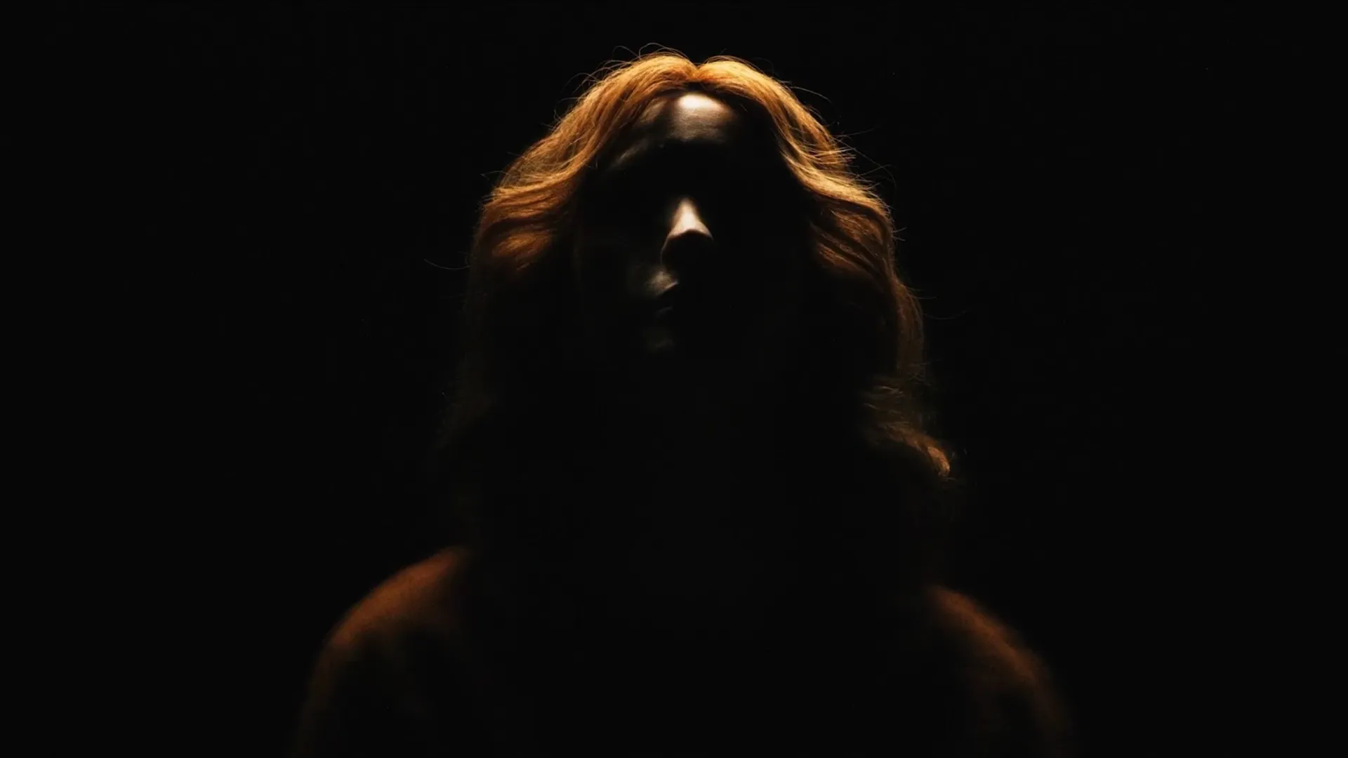 A figure with long copper hair stands in darkness, the face mostly in shadow, lit from above.