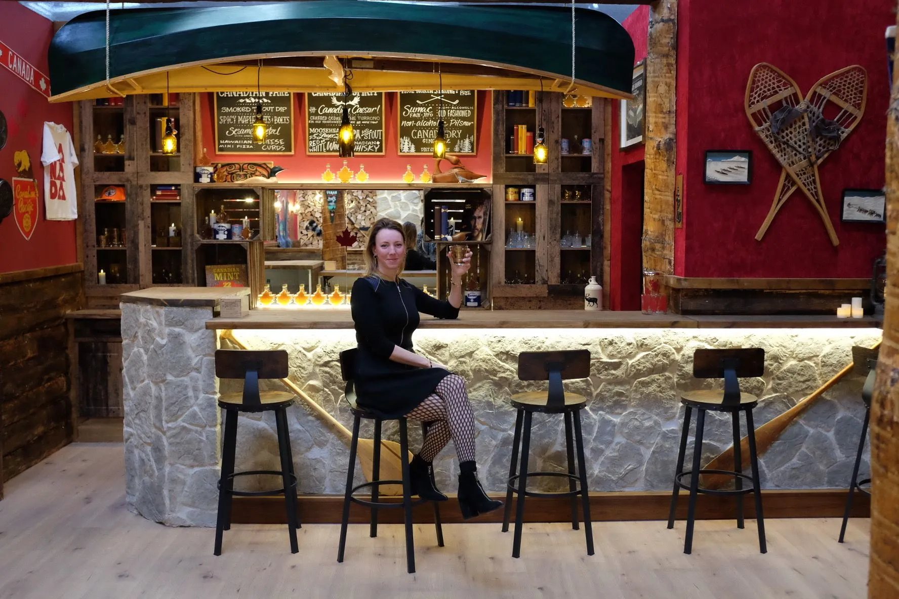 A guest raises a glass at a stone-fronted bar under a hanging green canoe, with chalkboard menus, maple syrup bottles and crossed snowshoes on a red wall.