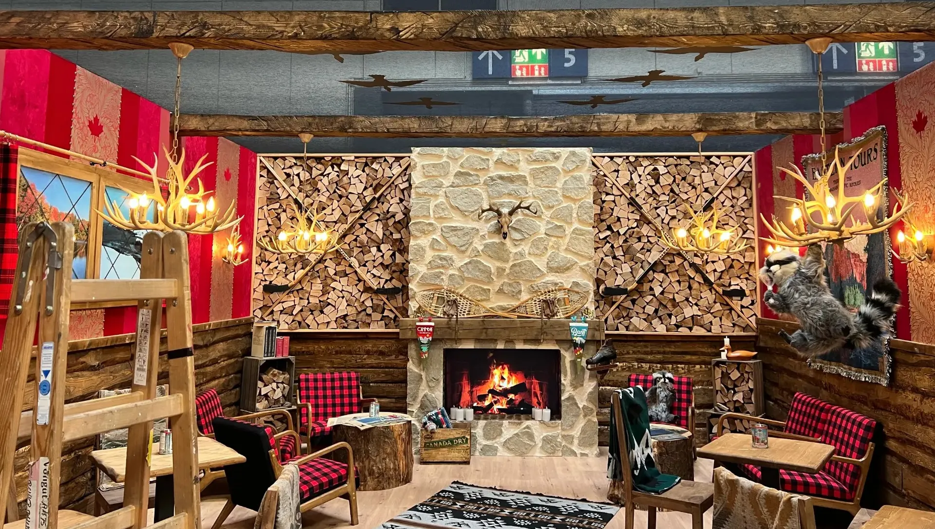 The full Canadian lodge: a stone fireplace between firewood walls with crossed hockey sticks, antler chandeliers, plaid armchairs and a raccoon swinging from a chandelier.