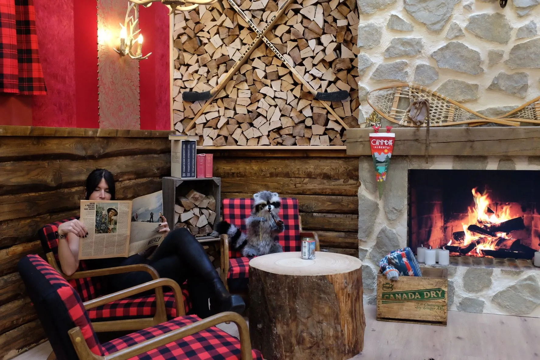 A Canadian lodge lounge: buffalo-plaid armchairs, a tree-stump table, a stacked-firewood wall with crossed hockey sticks, and a stone fireplace with snowshoes and a Canmore pennant.