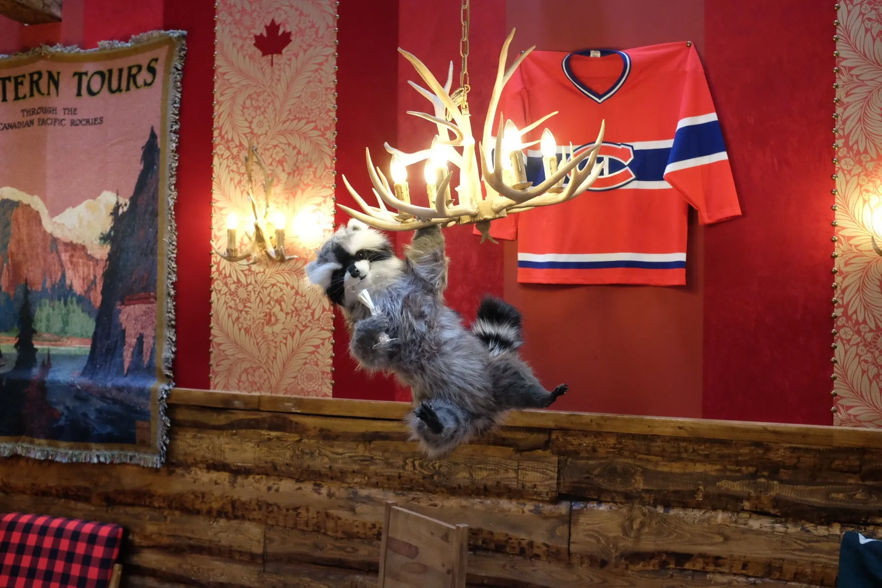A toy raccoon holding a cocktail glass swings from an antler chandelier in front of a Montreal Canadiens jersey and a vintage Rockies travel poster.