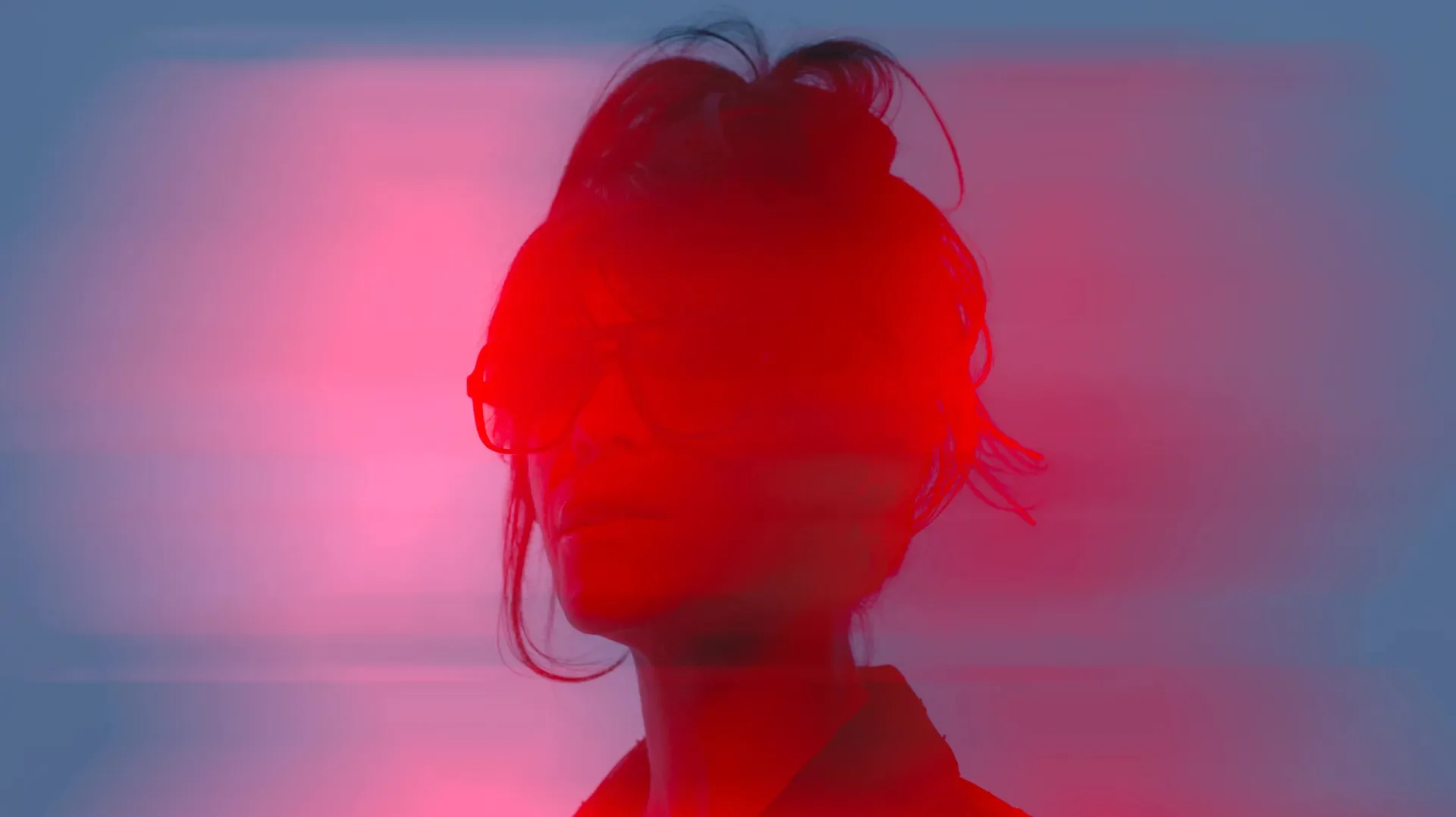 Close-up of a woman in sunglasses, her face washed in red light against a blurred blue background.
