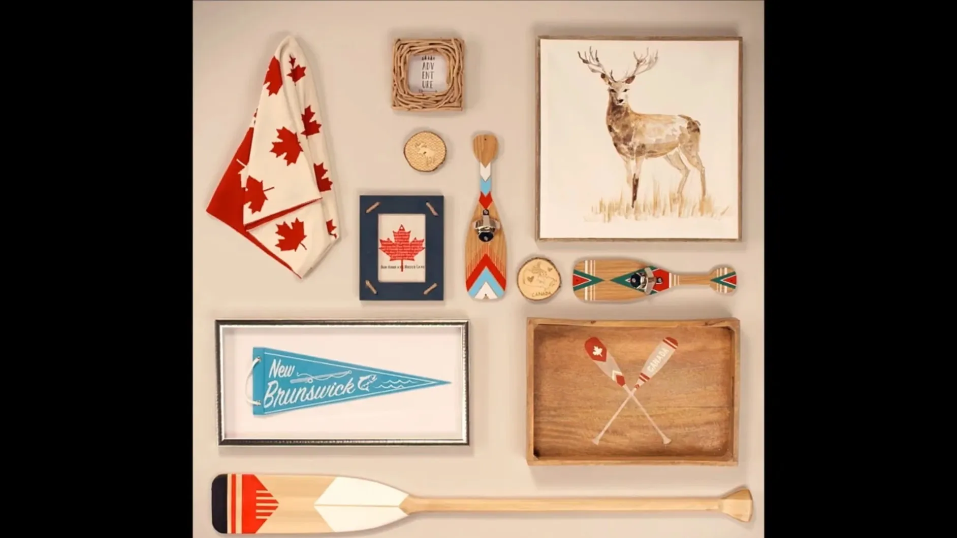 A flat lay of Canadiana decor: a maple-leaf throw, a painted deer, a New Brunswick pennant, crossed paddles on a wooden tray and a striped canoe paddle.