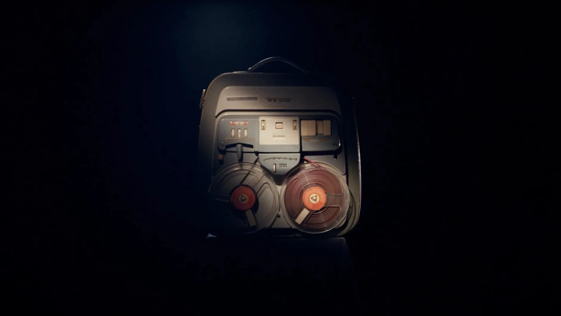A vintage reel-to-reel tape recorder in a carrying case, lit by a single spotlight in darkness.