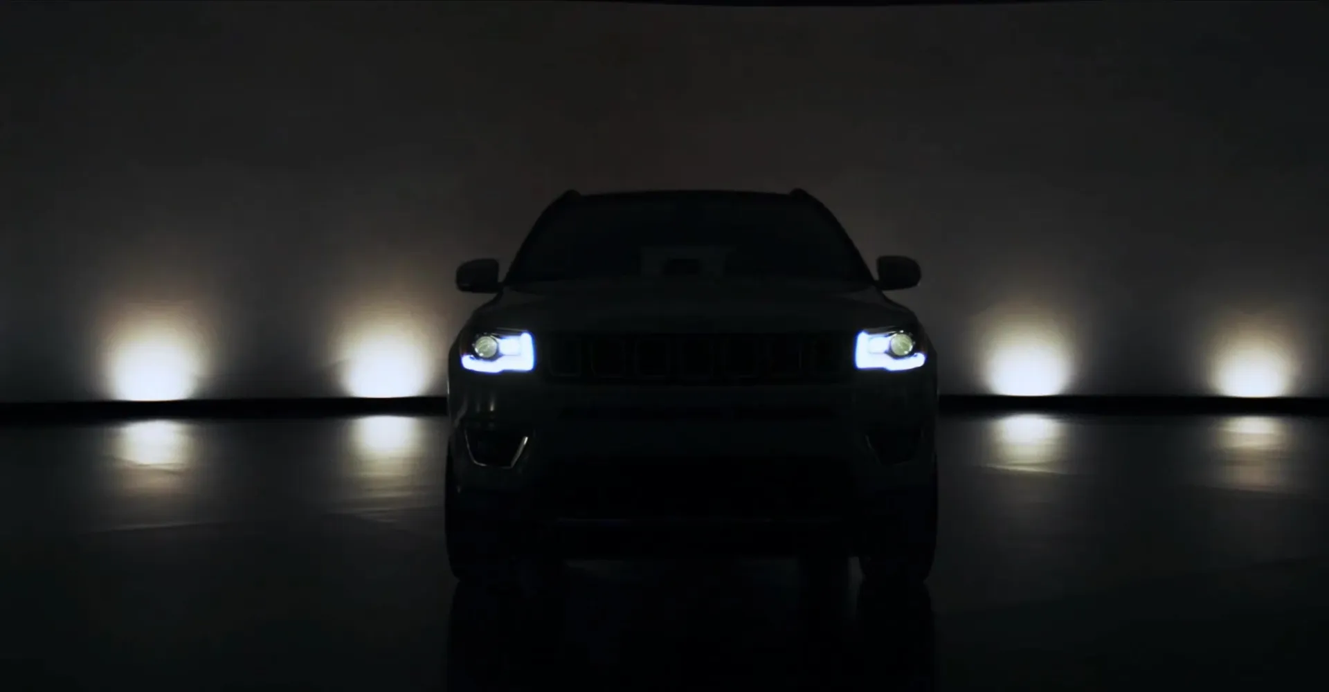 A Jeep in silhouette facing the camera, headlights on, in a dark studio lit by soft floor lights.