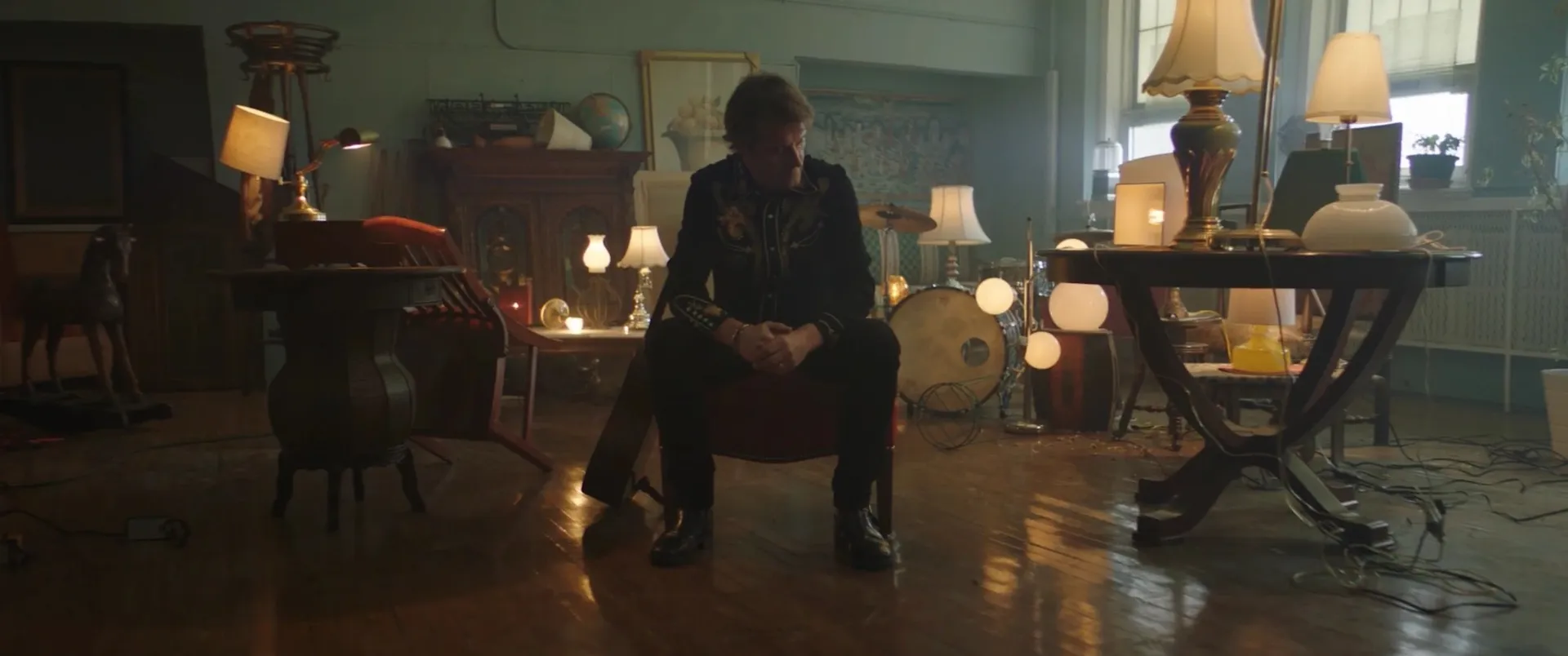 Jim Cuddy sits on a chair in a dim room crowded with glowing vintage lamps, antique furniture and a drum kit.