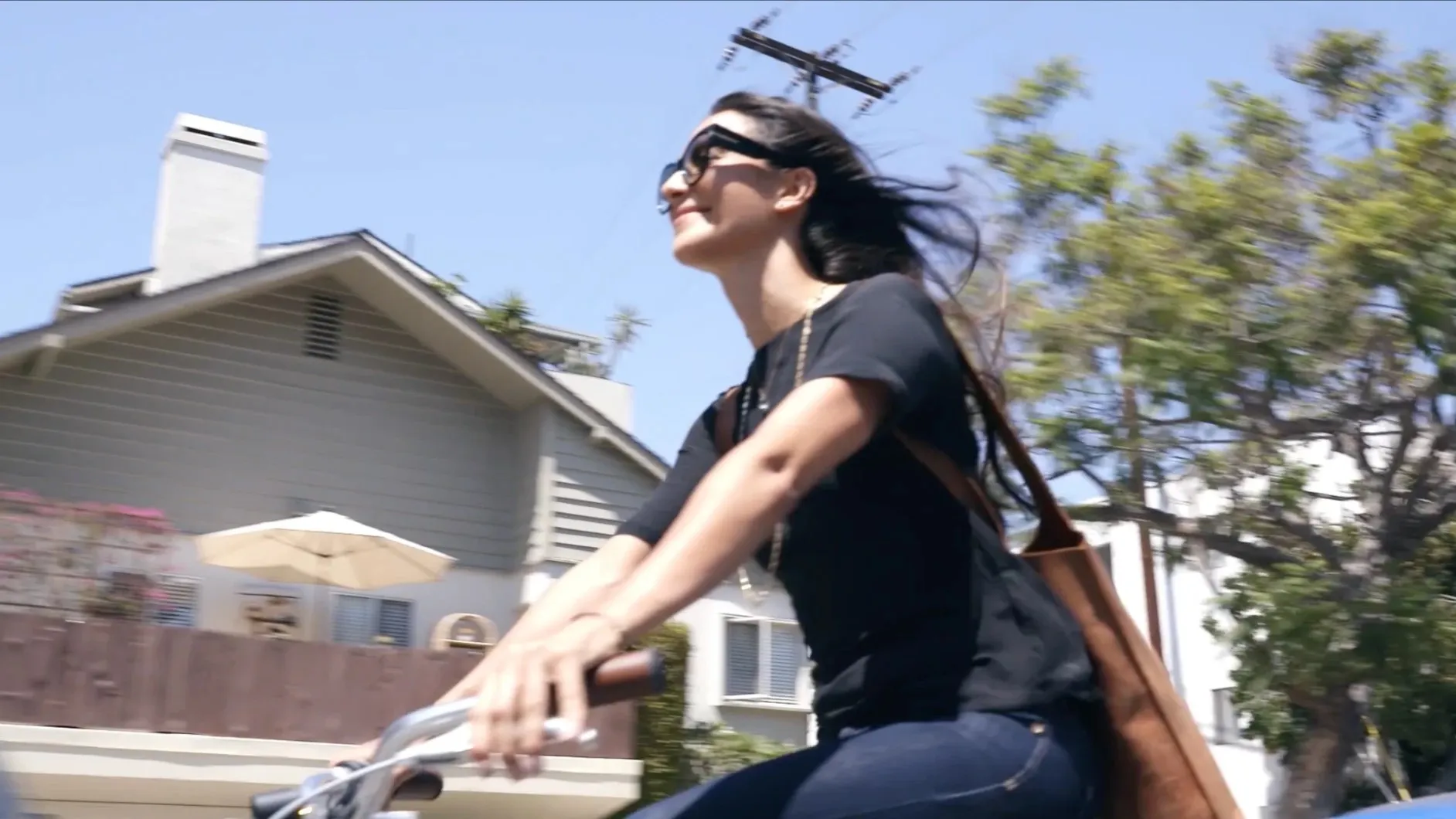 A smiling woman in sunglasses and a black tee rides a bike down a sunny residential street, hair flying, a leather bag over her shoulder.