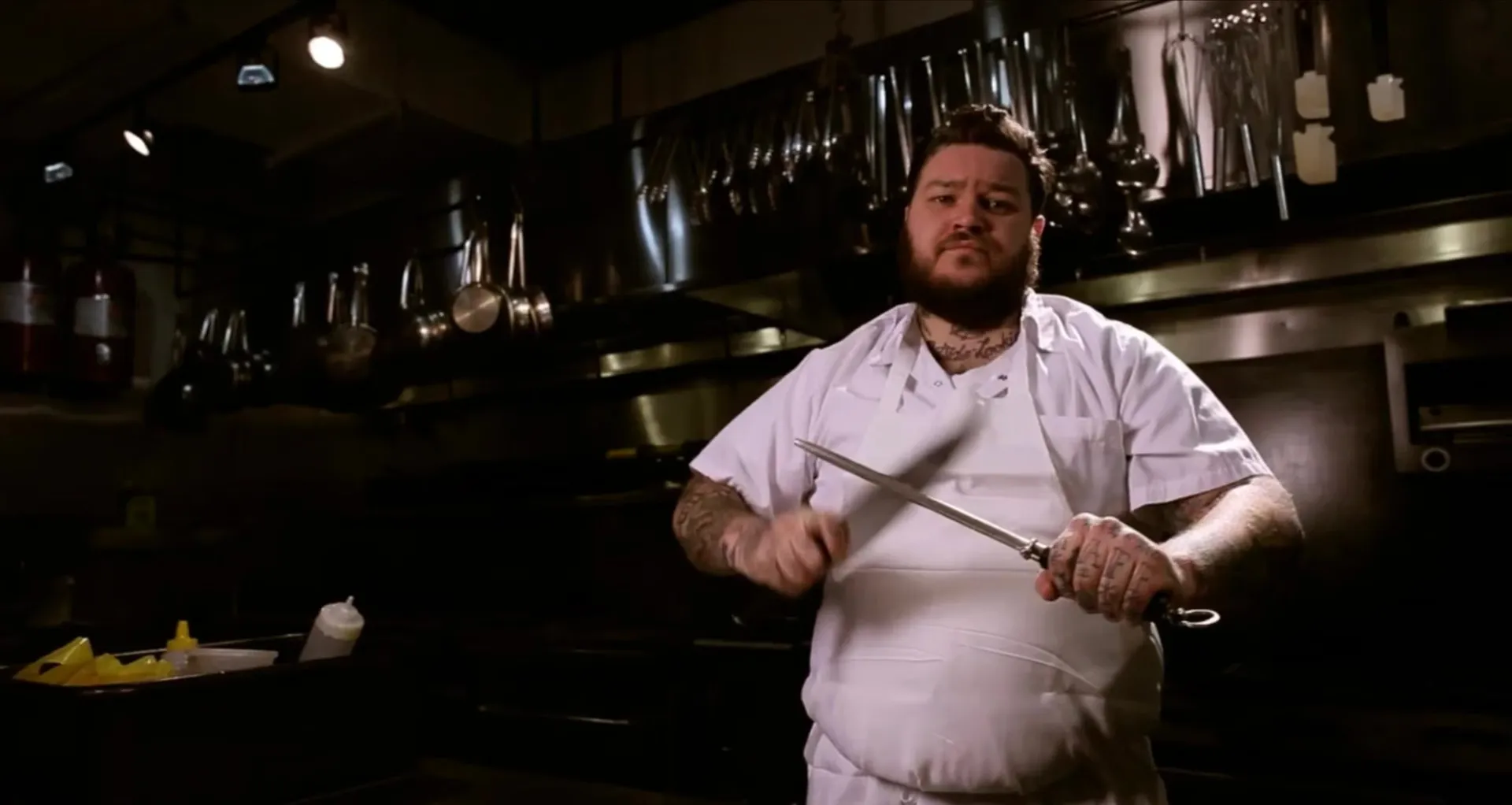 Matty Matheson in chef whites sharpening a knife on a steel in a dark restaurant kitchen.