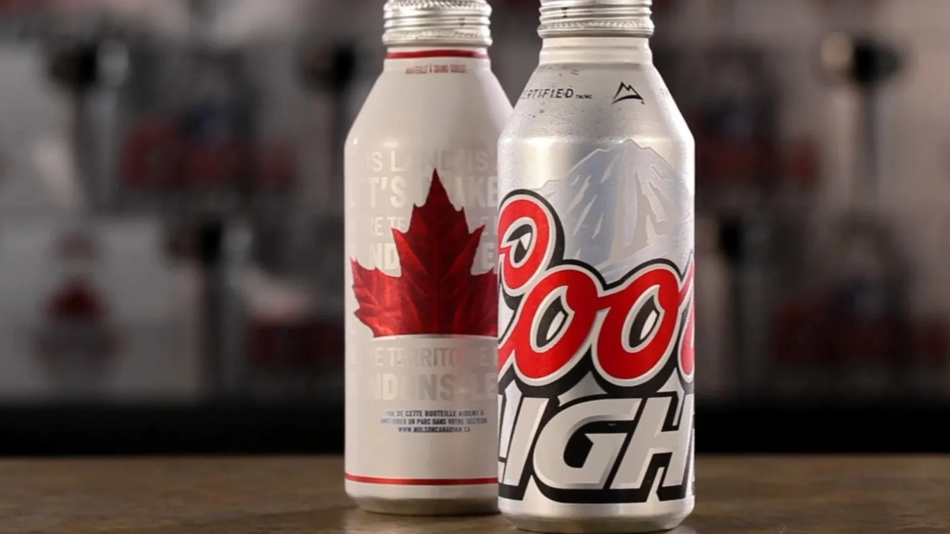 A Molson Canadian bottle with a red maple leaf beside a frosted Coors Light bottle on a bar top.
