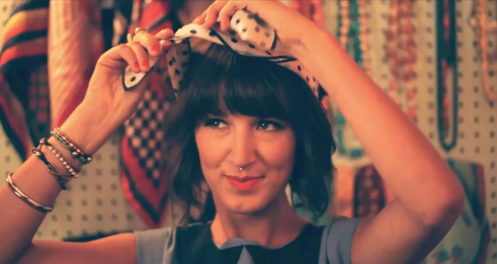 A smiling woman with a fringe ties a polka-dot scarf into her hair in front of a wall of vintage scarves and necklaces.