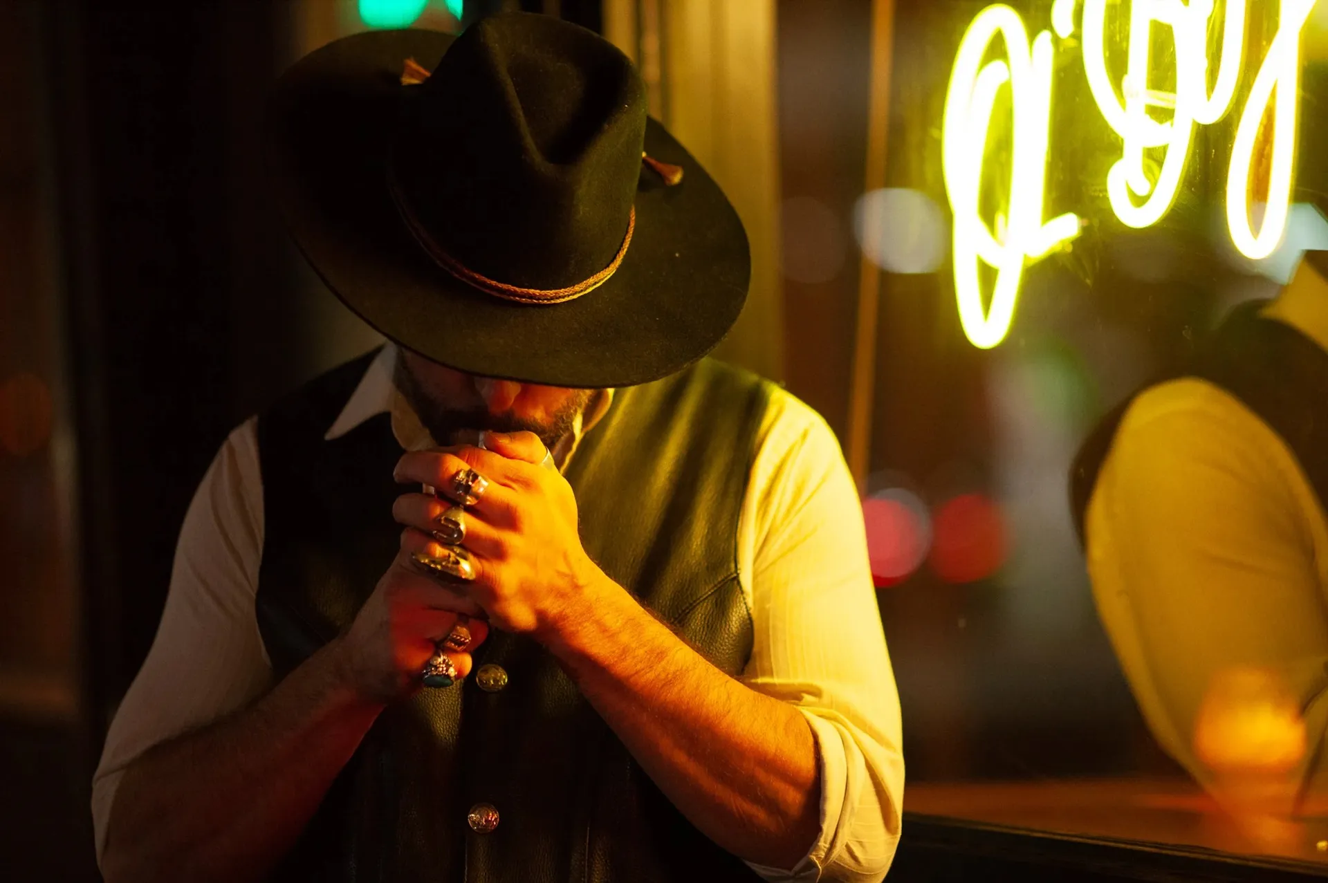 A man in a black hat and leather vest bows his head to light a cigarette, ringed hands cupped, beside the yellow Ol' Boy neon sign.