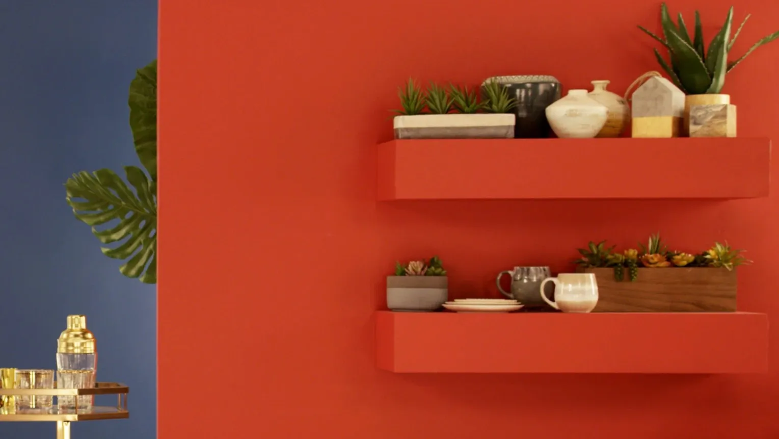 Floating red shelves of succulents, vases and mugs on a coral wall, beside a navy panel, a monstera leaf and a gold bar cart.