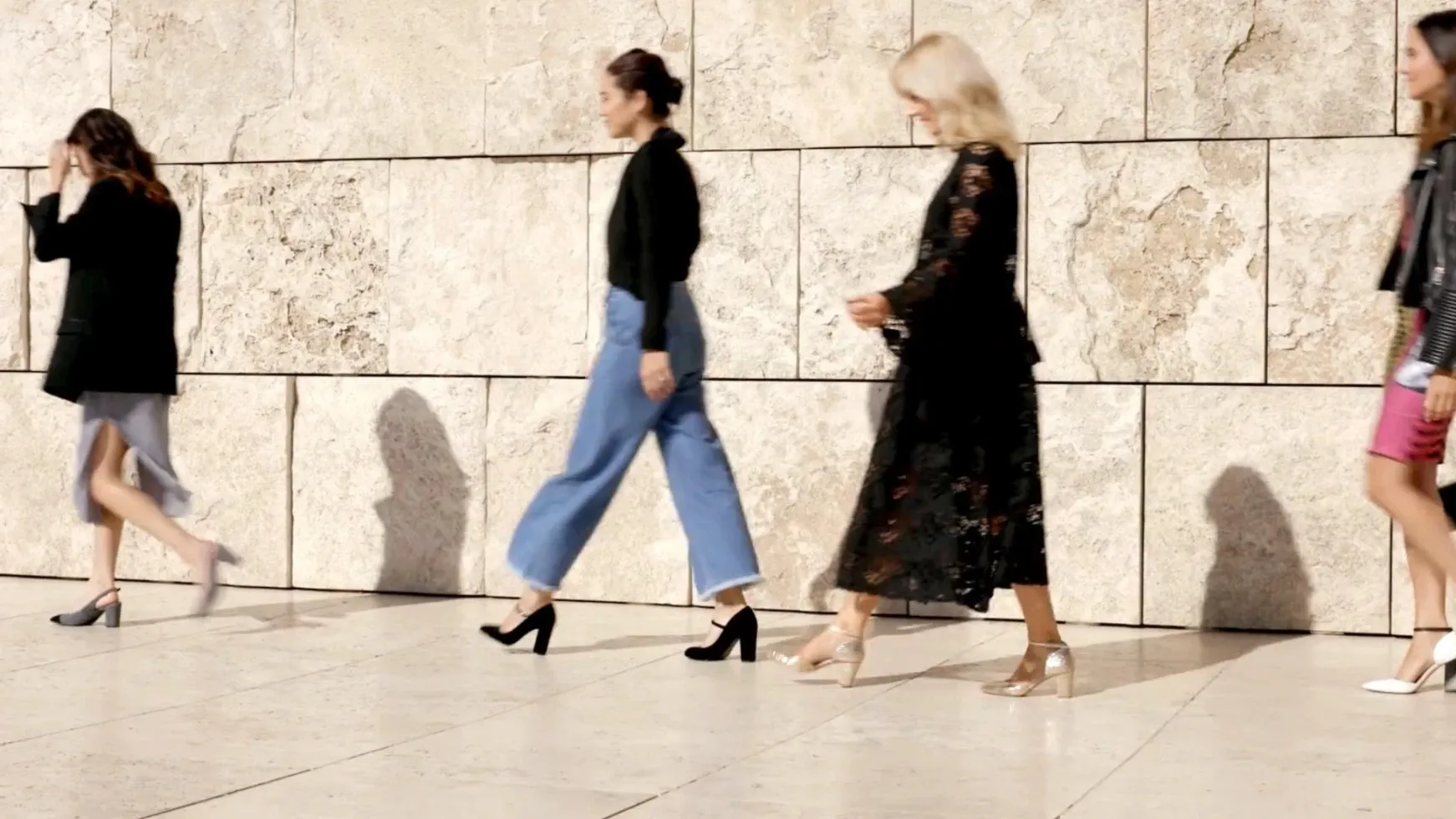 Four women in heels walk in a line past a sunlit travertine wall, their shadows cast beside them.