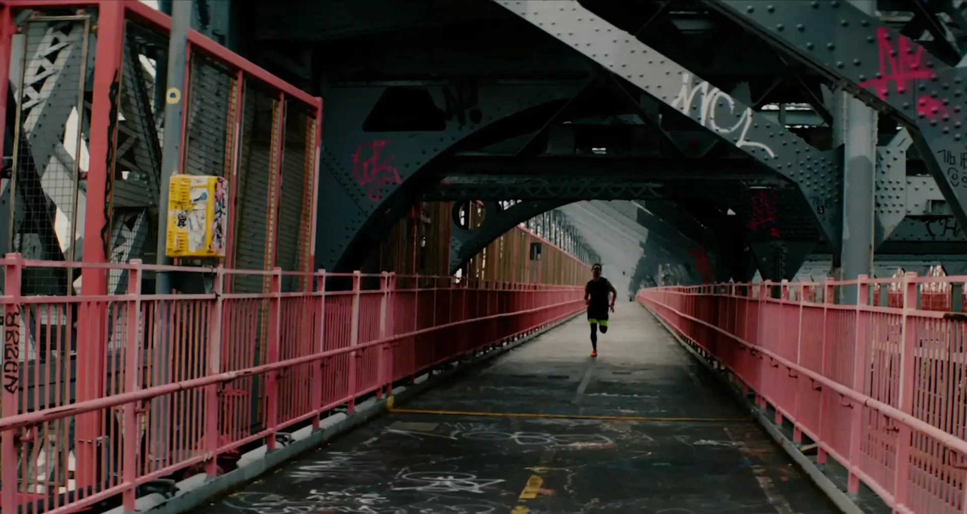 A runner sprints toward the camera along the pink-railed walkway of a steel bridge covered in graffiti.