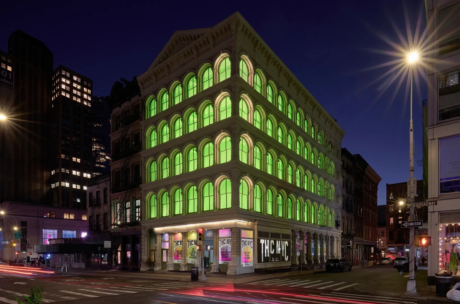 The SoHo cast-iron building at 427 Broadway at night, every arched window glowing green, THC NYC posters along the street front.