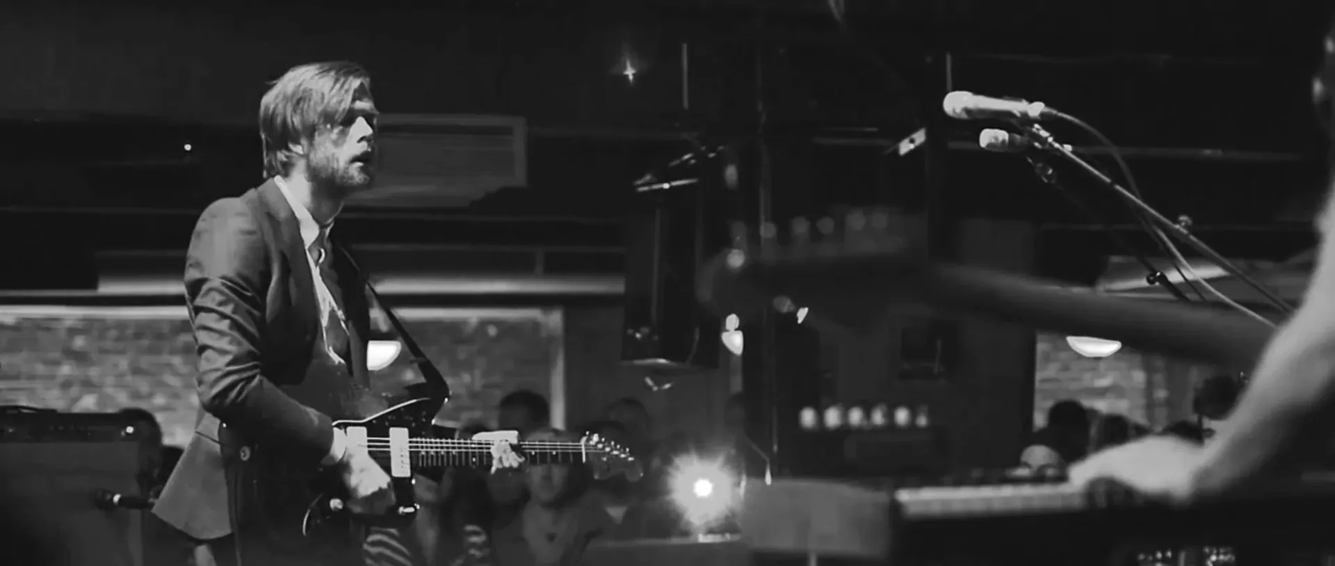 Black-and-white live shot of a guitarist in a suit playing on a club stage, the crowd behind him and a keyboard player blurred in the foreground.