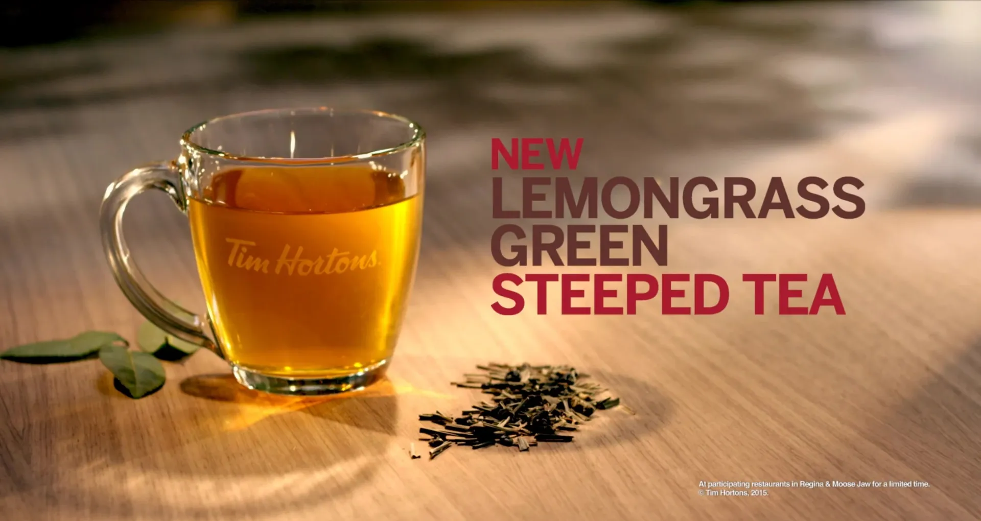 A glass Tim Hortons mug of tea on a wooden table beside loose lemongrass, with the words New Lemongrass Green Steeped Tea.