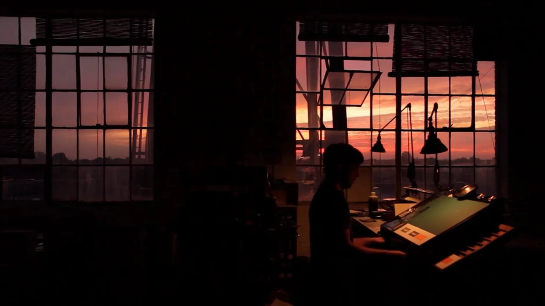A printer in silhouette works at a press in a dark studio, a pink and orange sunset glowing through tall factory windows.