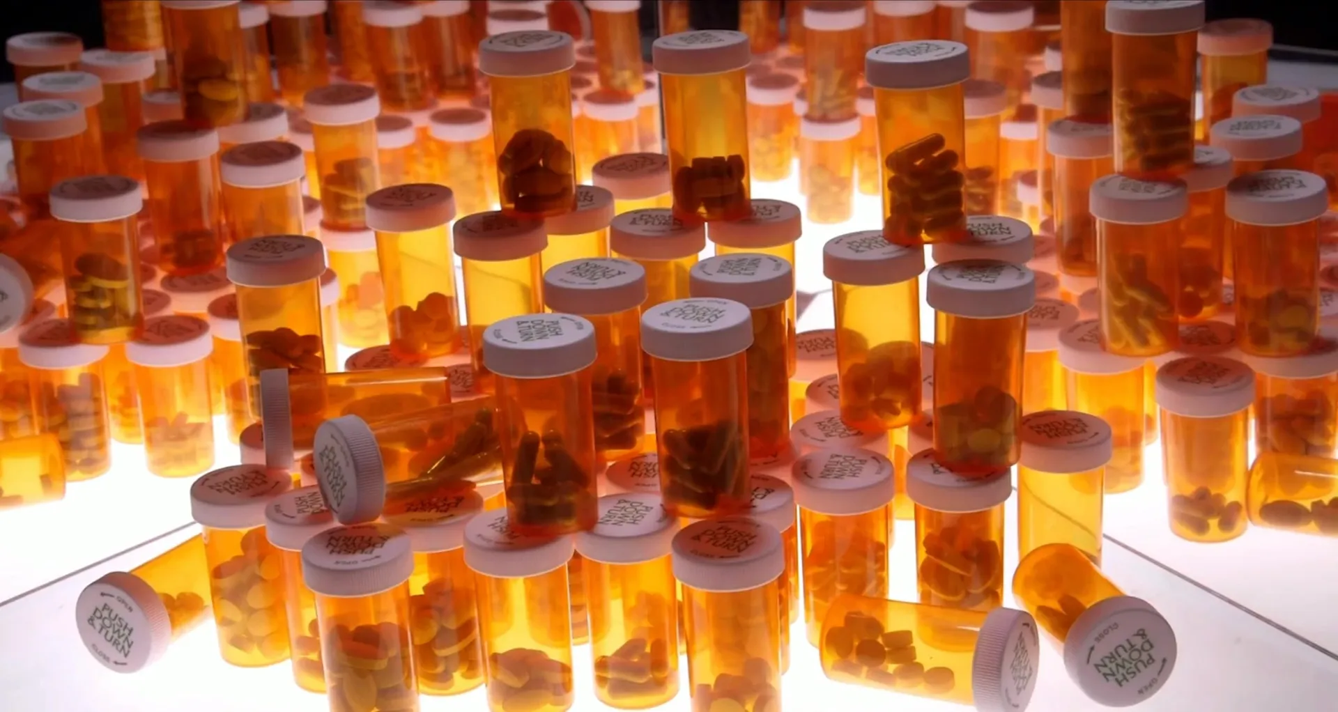 Hundreds of amber prescription bottles stacked on a glowing light table.