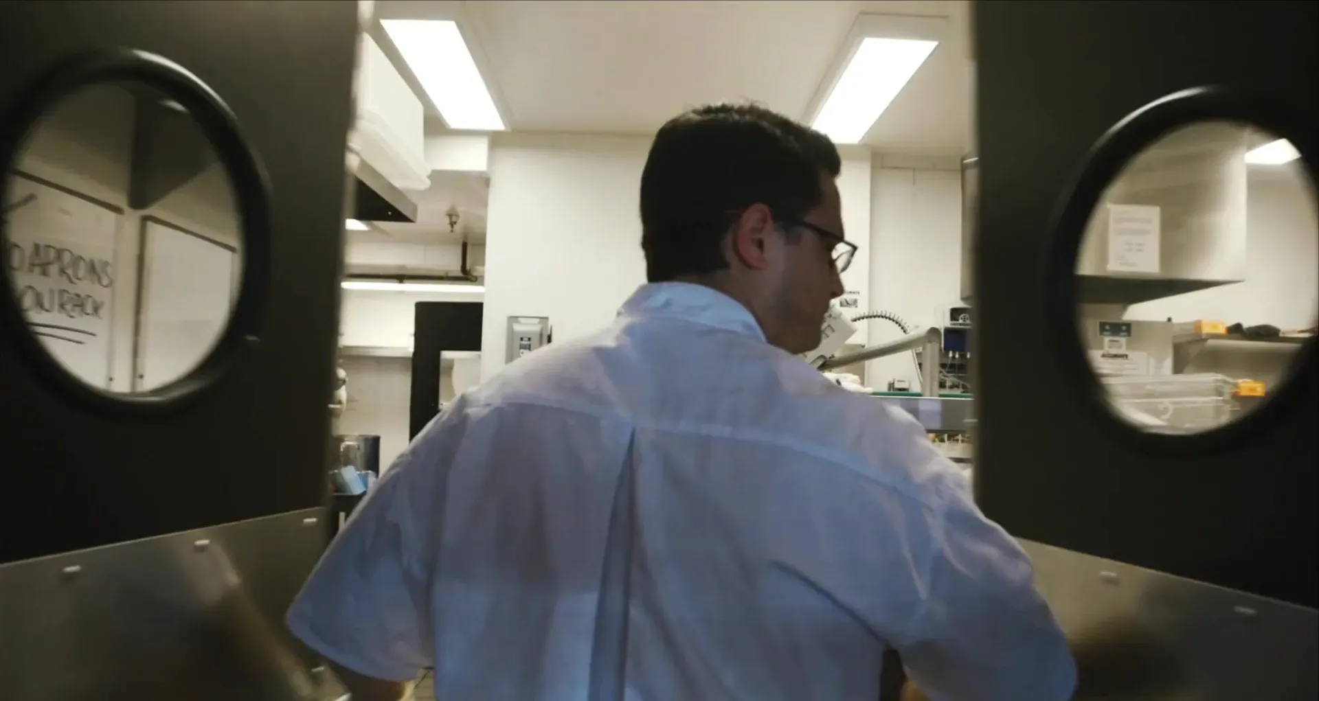 Seen through swinging kitchen doors, a chef in a white shirt works in a bright restaurant kitchen.